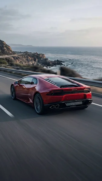 A cinematic 8-second shot of a red luxury sports car driving fast along a coastal highway. Ocean on the right, cliffs and rocky shoreline on the left. Golden hour light, shallow depth of field, smooth camera tracking.